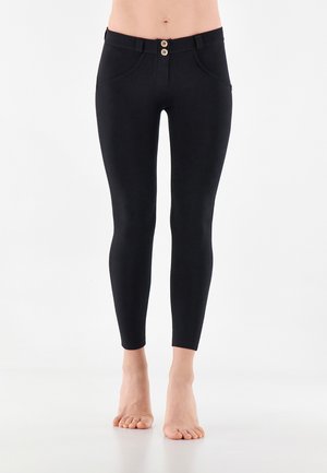 Mustat vartalonmyötäiset nilkkapituiset housut, joissa on kaksi puisia nappia vyötäröllä, jalaton seisovan henkilön yllä yksiväristä taustaa vasten.