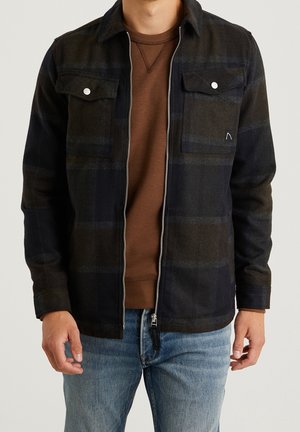 Homme portant une veste à carreaux noir et vert foncé non zippée sur un pull marron et un jean bleu clair, sur fond uni.