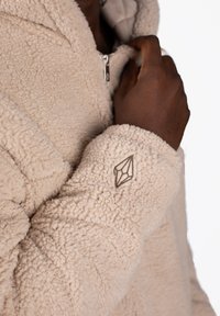 Cream-colored fleece jacket with a plush texture, featuring a zip front and a logo detail embroidered on the cuff.
