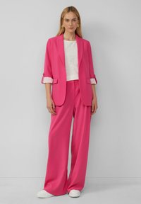 Fuchsia pink blazer with rolled sleeves, paired with matching wide-leg trousers and a white textured top, complemented by white sneakers.