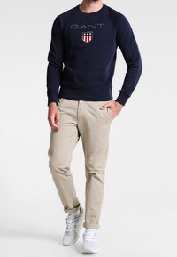 Felpa blu navy con logo "GANT" e design a scudo, abbinata a chinos beige e sneakers bianche. Tessuto liscio con polsini a coste.