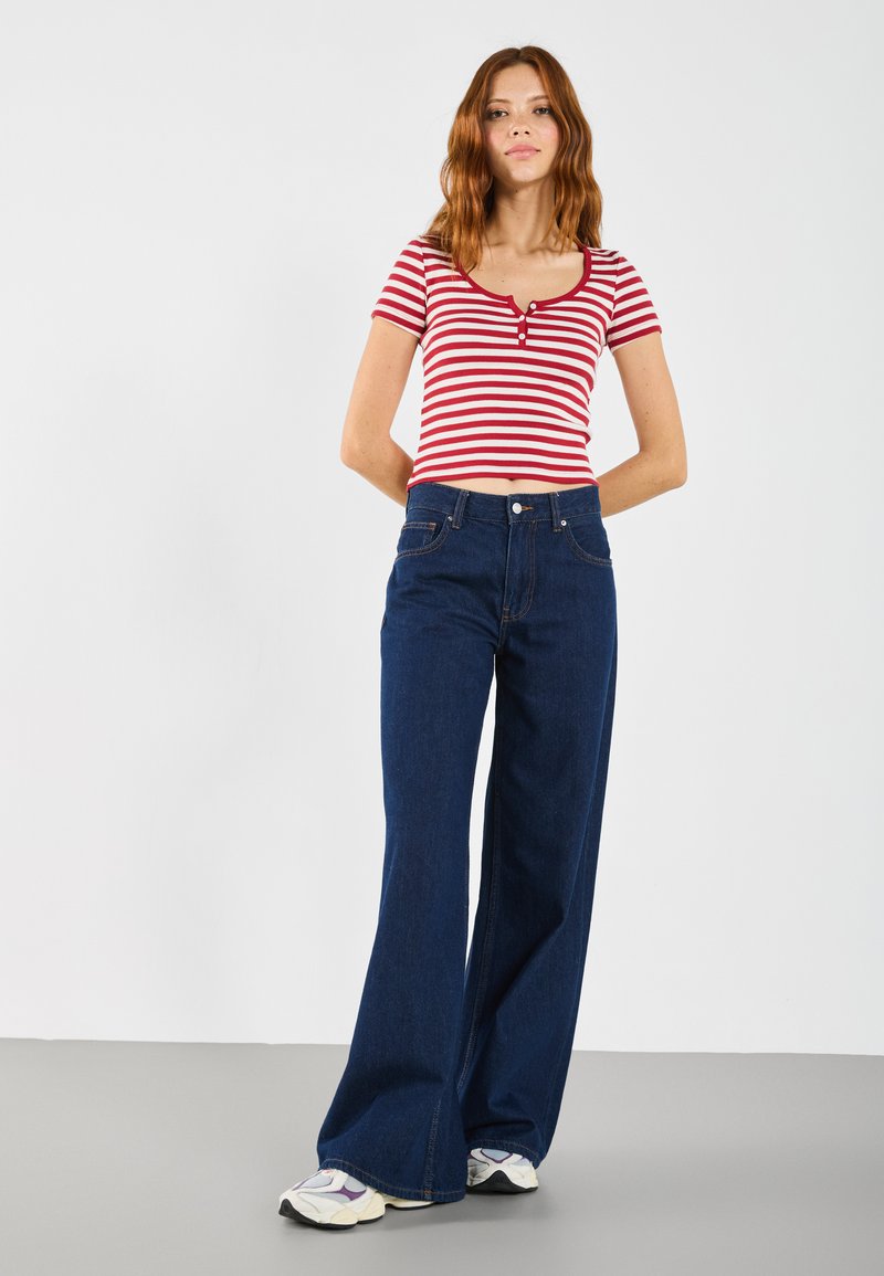 Camisa de rayas rojas y blancas de manga corta con detalle de botones en la parte delantera, combinada con unos vaqueros de pierna ancha de cintura alta en azul oscuro y zapatillas blancas.