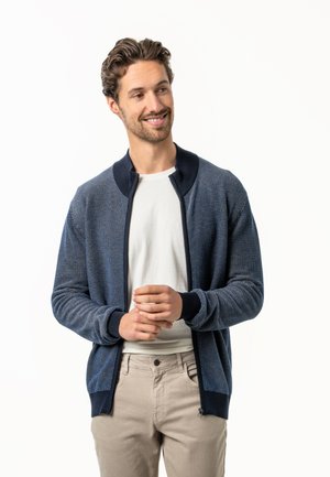 Smiling man wearing a navy blue patterned zip-up cardigan over a white shirt and beige pants, standing with hands loosely clasped.