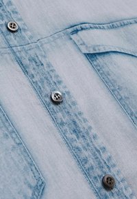 Camicia in denim azzurro chiaro con chiusura a bottoni, dettagli sulla tasca e bottoni scuri a contrasto, caratterizzata da un design in tessuto testurizzato.