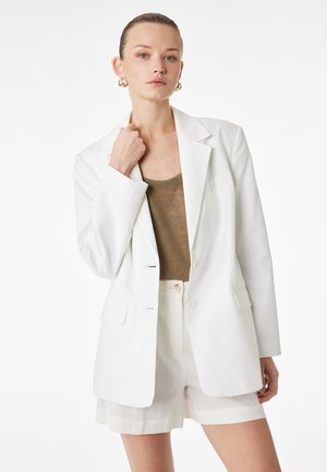 Femme aux cheveux plaqués en arrière portant des boucles d'oreilles créoles dorées, un blazer blanc, un top marron et un short blanc, posant avec une main près de son col.