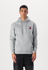Grå hoodie med en frontlomme, der har et lille logopatch i rød, hvid og blå. Fremstillet af et blødt, struktureret stof.