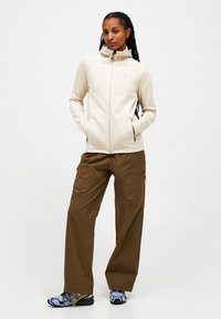 Világos bézs, cipzáros dzseki magas gallérral, varrás nélküli kialakítással és oldalsó zsebekkel, barna cargo nadrággal és több színben pompázó sportcipővel párosítva.