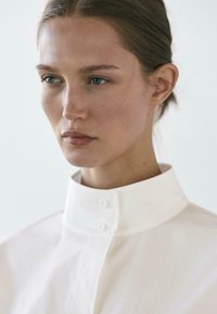 Witte blouse met kraag, gemaakt van gladde stof, met een hoge kraag en twee knopen aan de voorkant, die een minimalistisch ontwerp benadrukken.