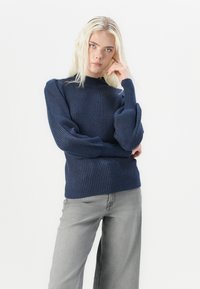Navy ribbed knit sweater with a high neck and puffed sleeves, paired with light grey wide-leg jeans, set against a plain white background.