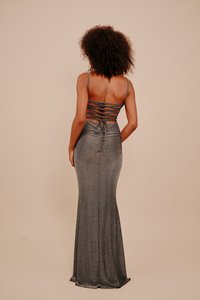 Ženska s kodrastimi lasmi, oblečena v oprijeto srebrno oblekico z odprtim hrbtom in križnimi naramnicami, stoji ob enostavni bež ozadju.