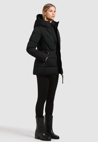 Kvinde iført en sort pufferjakke med hætte, sorte leggings og sorte støvler til midt på læggen, stående med den ene hånd i jakke kopi.