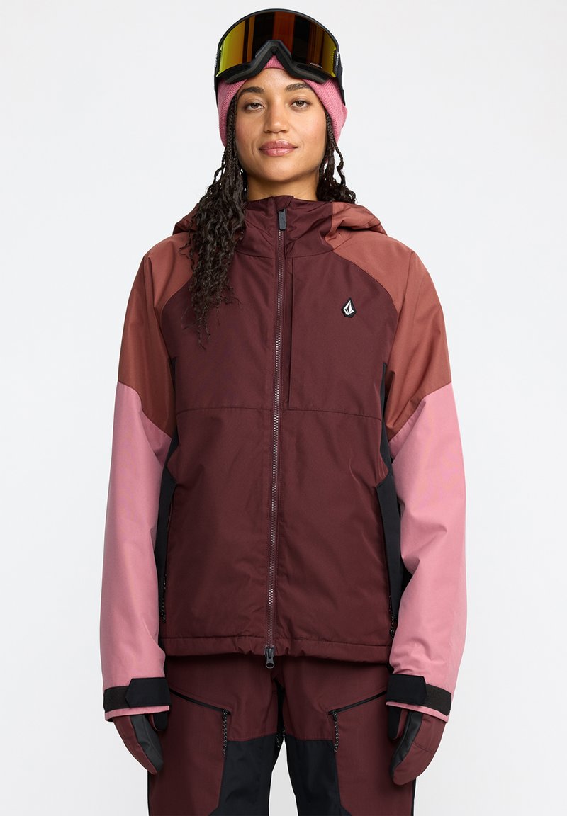 Veste imperméable bordeaux avec des accents roses et noirs. Elle possède un zip à l'avant, une capuche et des poignets renforcés. Portée avec des lunettes de ski et des gants.