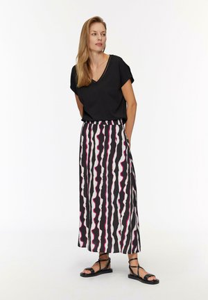 T-shirt noir à col en V associé à une jupe longue avec des rayures ondulées noires, blanches et roses alternées. Des sandales plates noires complètent le look.