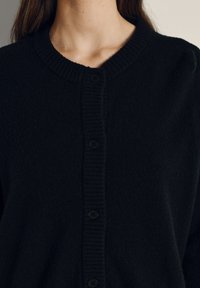 Črna jopica iz mehkega pletenega materiala, z okroglim izrezom, zaključki z rebrastim vzorcem in šestimi črnimi gumbi spredaj.