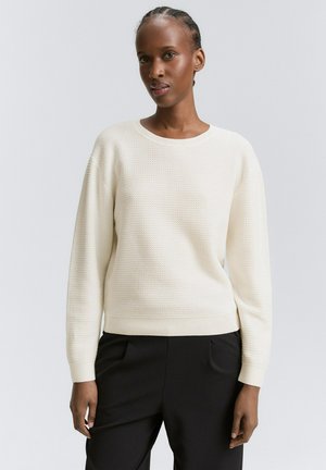 Pull texture beige clair avec un col rond et des manches longues, doté de poignets et d'un ourlet côtelés. Associé à un pantalon noir.