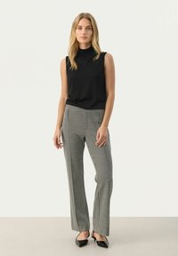 Zwarte mouwloze top met hoge hals, gecombineerd met broek met een ruitpatroon. Model draagt puntige zwarte hakken. Gladde stofstructuur.