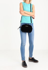 Žena v tyrkysovém bezrukávovém topu, světlých modrých džínách slim fit, černých lakovaných botách a černé prošívané kabelce přes rameno, stojící se založenýma rukama.