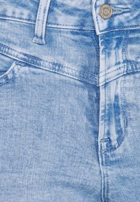 Mezclilla azul claro con una textura desgastada, presenta una cinturilla, pasadores para el cinturón y costuras cosidas. Incluye un detalle de botón metálico.