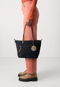 Marinblå tygväska med bruna detaljer, dragkedjeficka och dekorativ nyckelring. Bärs med rosa byxor och bruna snörskor.