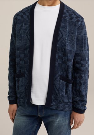 Homme portant un cardigan bleu marine texturé à ouverture devant avec des motifs géométriques, par-dessus un t-shirt blanc et un jean bleu, debout devant un fond neutre.