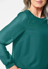 Türkise Bluse mit langen, transparenten Ärmeln und einem runden Ausschnitt. Verfügt über eine dezente Textur und eine kleine Tasche auf der linken Seite.