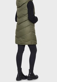 Olive green puffer vest with a hood, featuring a chevron quilted design. Worn over a black long-sleeve top and paired with black skinny jeans and boots.