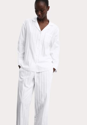 Donna che indossa una camicia da pigiama bianca larga con bottoni e pantaloni largi coordinati, in piedi con le mani nelle tasche davanti a uno sfondo semplice.