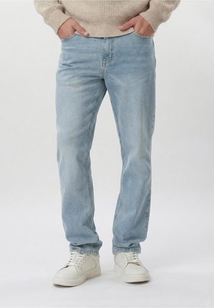 Homme portant un jean bleu clair, un pull beige et des baskets blanches, les mains dans les poches avant, face à un fond uni.