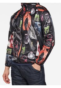 Veste noire à fermeture éclair avec une finition brillante et des motifs imprimés colorés représentant divers objets, notamment des lunettes et des outils, sur le tissu.
