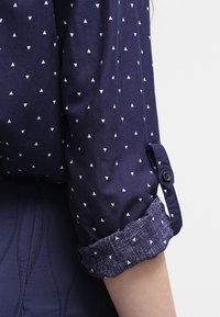 Chemise bleu marine avec motifs triangulaires blancs, présentant des poignets retroussés avec un accent bleu texturé. Le matériau semble doux et léger.