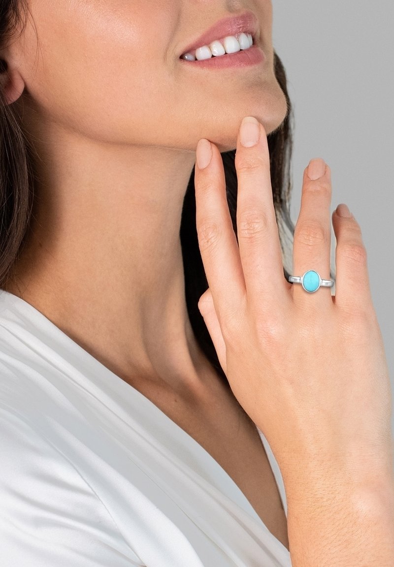 Bague en turquoise de forme ovale montée sur un anneau en argent. La main repose contre le visage avec un vernis à ongles neutre.