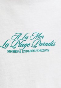 Τυρκουάζ κείμενο σε λευκό ύφασμα γράφει: «A La Mer La Plage Paradis» με καλλιγραφία και «SHORES & ENDLESS HORIZONS» με κεφαλαία γράμματα.