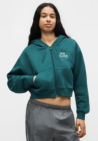 Jeune femme aux longs cheveux bouclés portant un sweat à capuche court zippé bleu sarcelle et un pantalon de sport gris sur un fond uni.