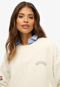 Roomkleurige sweatshirt met "SUPERDRY" logo, geribbelde halslijn en lange mouwen. Gedragen over een blauw-roze gestreept overhemd met kraag.