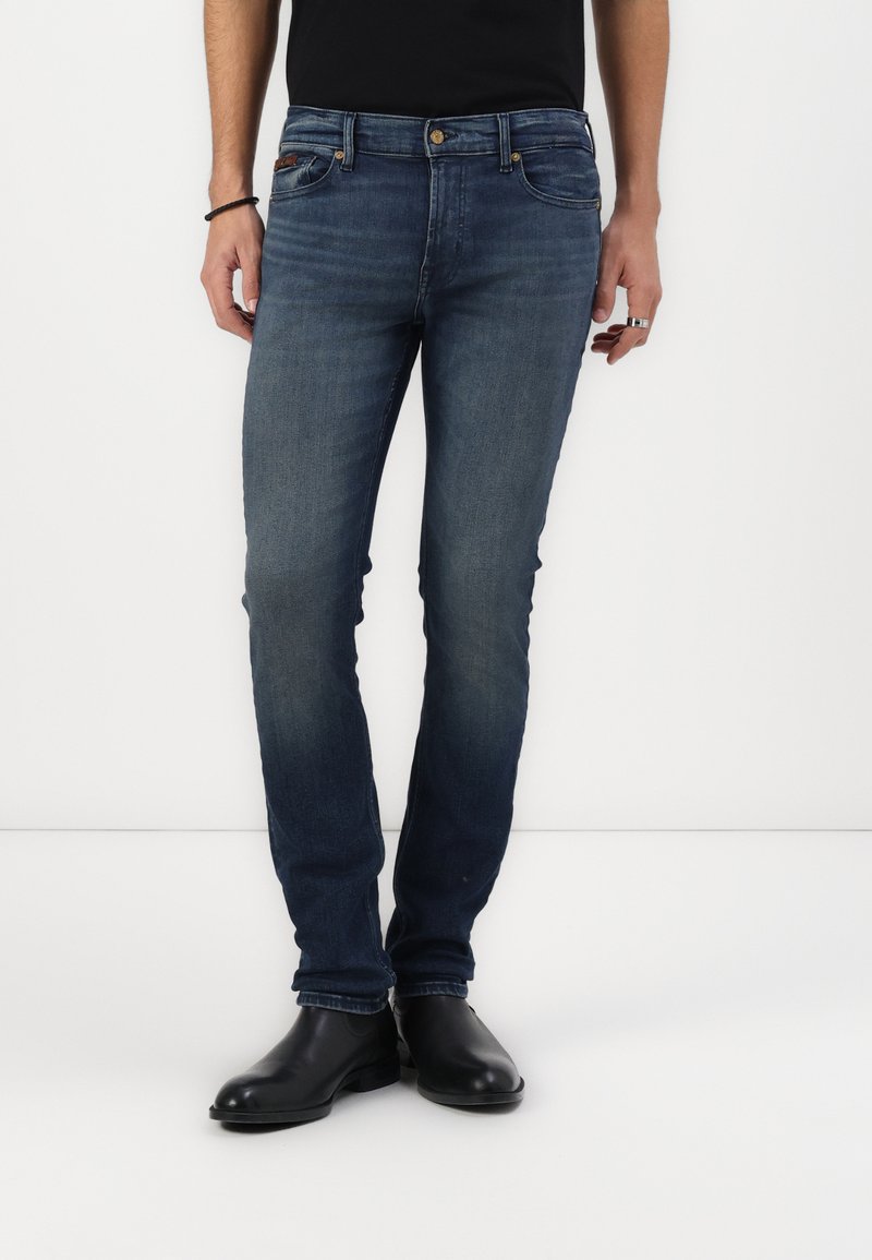 Jeans de mezclilla ajustados de color azul oscuro con un ligero desgaste, diseño de cinco bolsillos y herrajes metálicos. Llevados con botas negras hasta el tobillo.