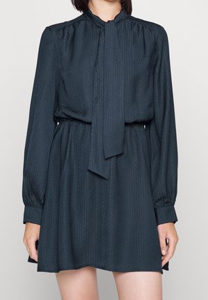 Femme portant une robe longue texturée sombre à manches longues, avec une taille froncée et un détail de cravate pendante à l'avant.