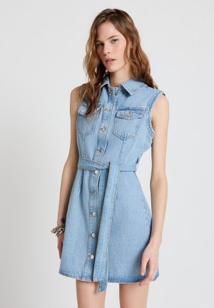 Junge Frau trägt ein ärmelloses hellblaues Jeanskleid mit Knöpfen vorne und passendem Gürtel, steht vor einem schlichten Hintergrund.
