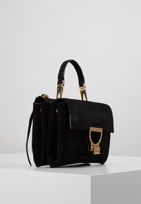 Mustan sametin käsilaukku, jossa on rakenteellinen muoto, kultaiset metalliosat, ylhäällä oleva kahva ja irrotettava olkahihna. Varustettu läppäsulkimella.