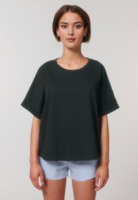 Een jonge vrouw staat zelfverzekerd tegen een eenvoudige lichte achtergrond, gekleed in een donkergroen oversized t-shirt en lichtblauwe denimshorts.