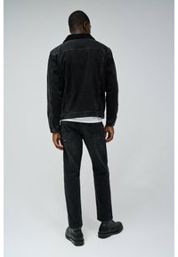 Chaqueta de corduroy negra con cuello de borrego, combinada con jeans oscuros y botas negras. Textura suave, forma ajustada y detalles mínimos.