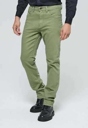 Jeans vert olive à coupe ajustée, cinq poches, et détails de surpiqûres visibles, assortis à des bottes noires à lacets.