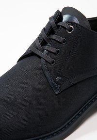 Chaussures en tissu noir avec une texture en chevrons, dotées de lacets plats, d'œillets ronds et d'un accent lisse près de la cheville.
