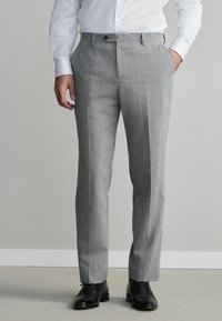 Pantalon taillé gris clair avec une texture subtile, plis avant, passants de ceinture et une coupe plate. Associé à des chaussures en cuir noires.