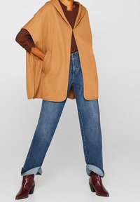 Persona con un poncho camel con bolsillos, una camiseta larga marrón, jeans azules arremangados y botas de tacón burdeos, de pie con las piernas abiertas.