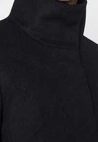 Černý kabát s vysokým límcem vyrobený z měkké, strukturované tkaniny. Má bezešvý design s minimálním šitím a štíhlou siluetou.