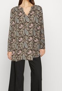 Túnica de mangas largas en tela marrón con un patrón floral de paisley en azul claro y rosa. Cuello en V y corte relajado. Combinada con pantalones negros.