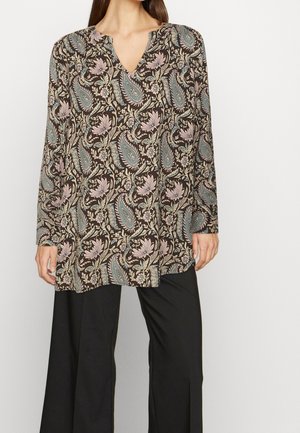 Langärmlige Tunika aus braunem Stoff mit einem paisley-blumigen Muster in Hellblau und Pink. V-Ausschnitt und lockere Passform. Kombiniert mit schwarzen Hosen.