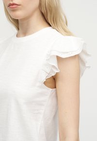 Ξανθιά γυναίκα που φοράει λευκό αμάνικο τοπ με βολάν και διακοσμητική δαντέλα τύπου eyelet γύρω από τις μασχάλες.