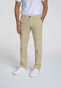 Chinos beige avec une coupe slim, fermeture à bouton et zip, poches latérales et une texture lisse, associés à des baskets blanches.