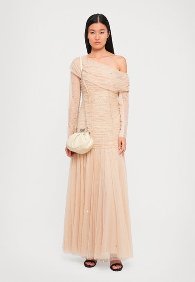 Vrouw in een vloerlange, off-shoulder beige jurk met pailletten, met een crèmekleurige handtas met een kettingriem en zwarteopen teen sandalen.